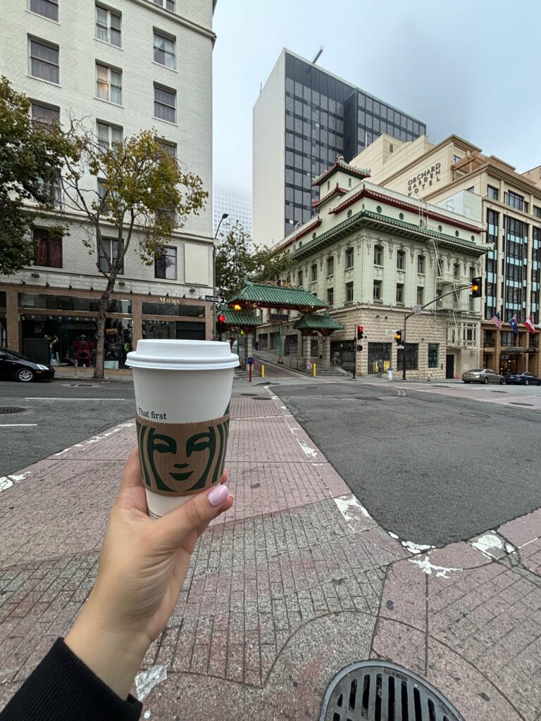 Tazza di caffè Starbucks con il logo della sirena tenuta in mano davanti al downtown di San Francisco con edifici storici e moderni