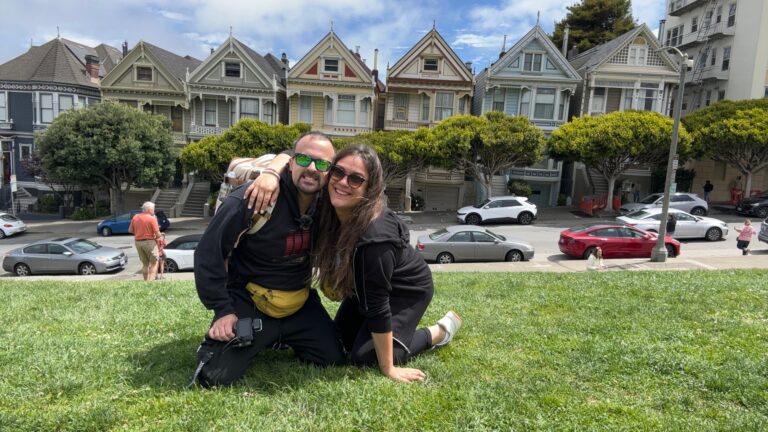 Alessia e Michael di Red Lace abbracciati su prato con dietro le case vittoriane colorate delle Painted Ladies di Alamo Square Park a San Francisco