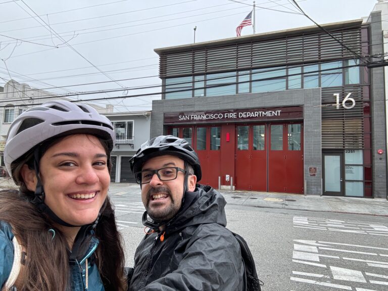 Alessia e Michael sorridono indossando caschi da vigile del fuoco davanti alla caserma San Francisco Fire Department Station 16 con porta rossa e bandiera americana