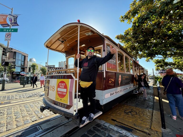 Michael di Red Lace sorride con la mano alzata sul Cable Car numero 28 rosso e marrone di San Francisco, con altri passeggeri sullo sfondo e foglie di alberi verdi sopra