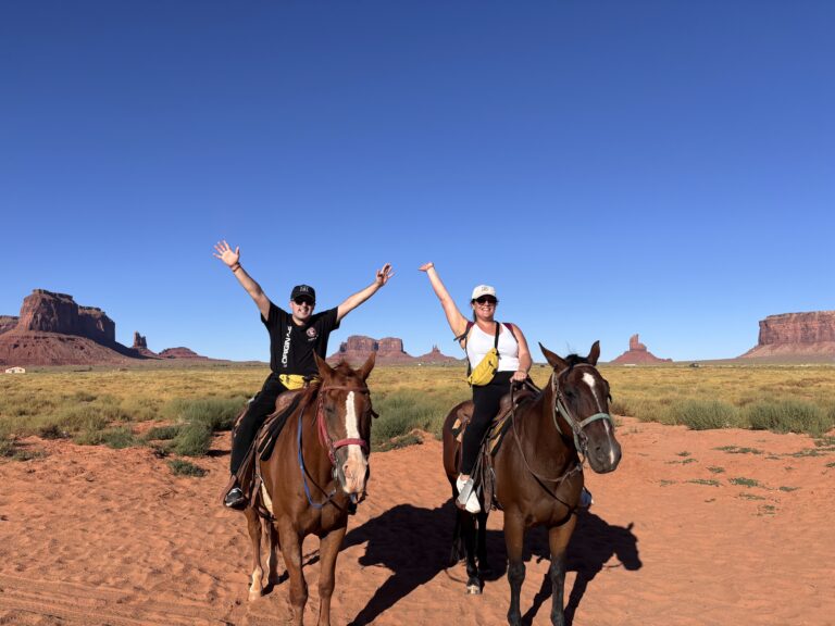 Alessia e Michael a cavallo con braccia alzate in Monument Valley con buttes rossi sullo sfondo