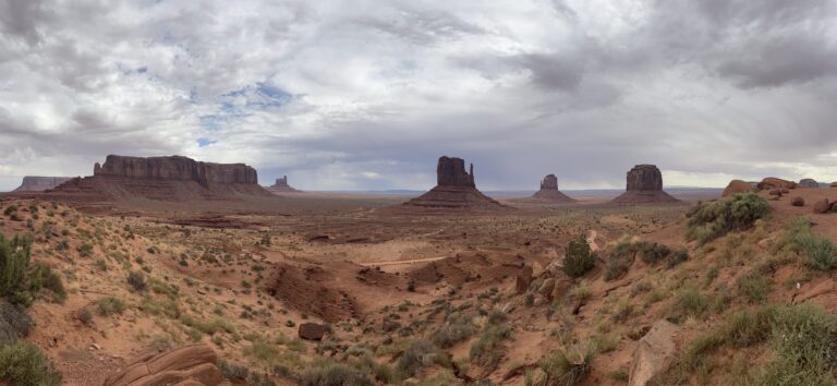Le tre Mitten Buttes di Monument Valley con vegetazione verde nel primo piano e cielo nuvoloso