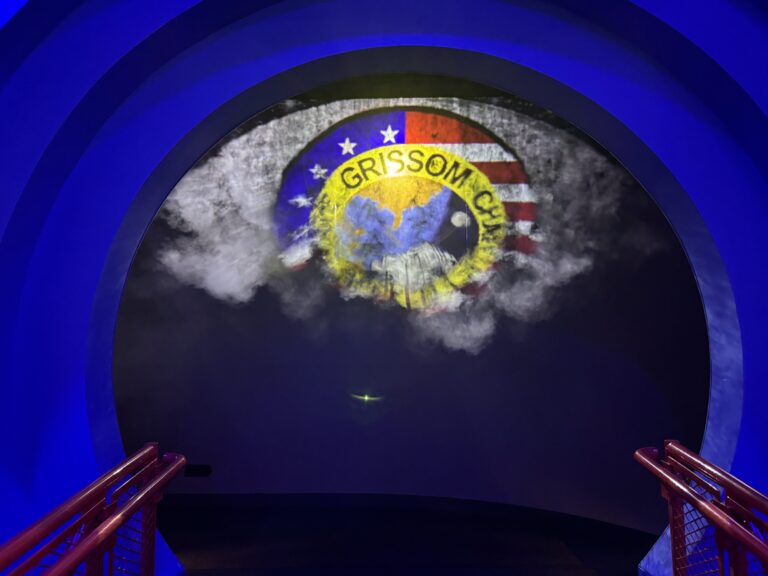 Stemma della missione Apollo 1 con il nome della missione scritto in giallo, circondato dalla bandiera americana, proiettato su una parete illuminata in blu al Kennedy Space Center in Florida - Memoriale dedicato alla missione tragica del 1967