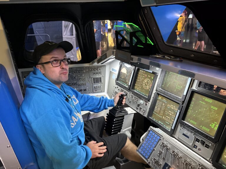 Michael di Red Lace posa dentro la replica della cabina di pilotaggio dello Space Shuttle Atlantis, indossando una giacca blu Kennedy Space Center, accanto ai comandi e pannelli di controllo della navetta