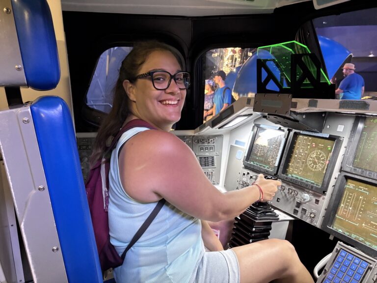Alessia di Red Lace posa sorridendo dentro la replica della cabina di pilotaggio dello Space Shuttle Atlantis al Kennedy Space Center in Florida, accanto ai comandi e ai pannelli di controllo della navetta