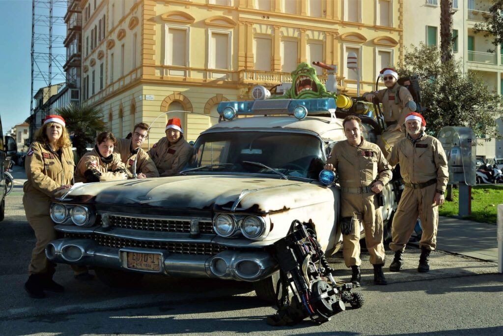 Gilles e l'intera squadra in uniforme da Ghostbusters posano orgogliosi davanti al muso della Ecto-1 Italiana finita, con Slimer visibile sul tetto.